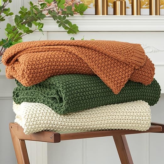 Chunky Cable Knit Throw Blankets stacked on top of each other. One orange, one green, and one beige.