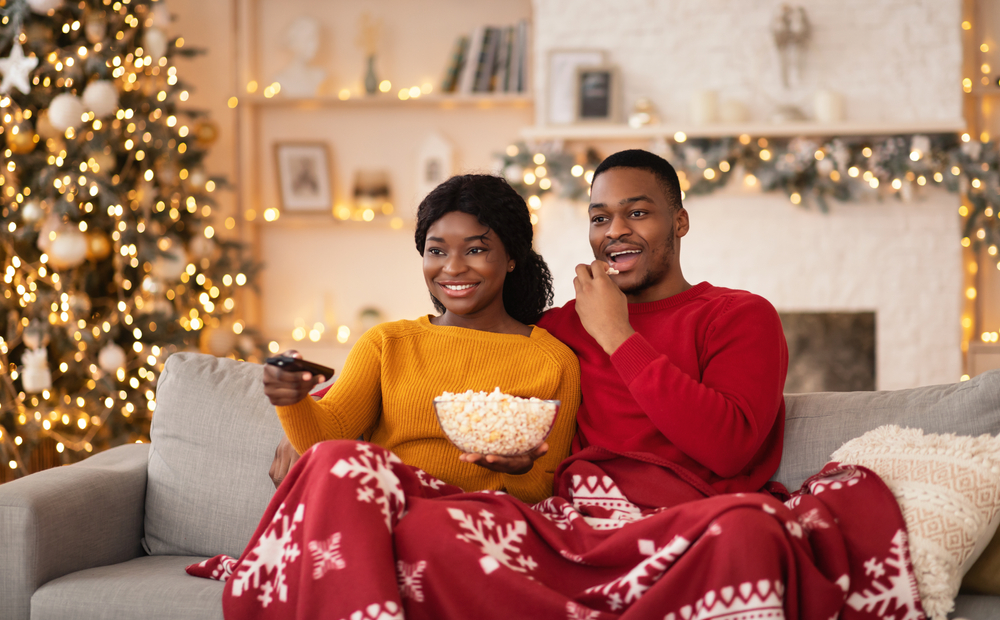 husband and wife with remote control and popcorn covered with blanket and watching tv in interior with tree