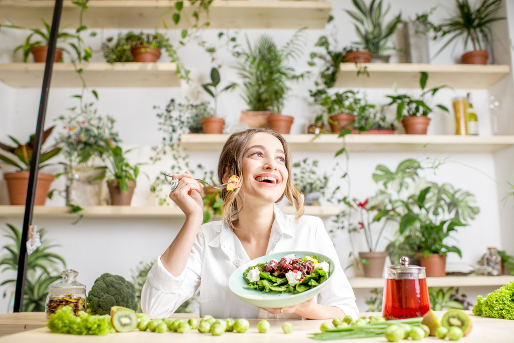 Young woman eating healthy food sitting in the beautiful interior with green flowers on the background