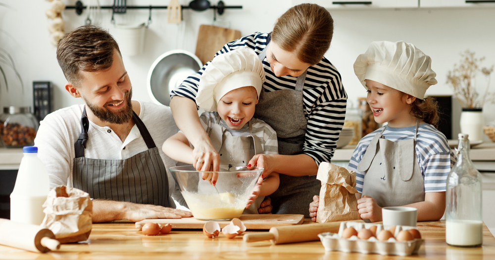 Happy young family with kids gathering in kitchen and preparing dough for pastry while spending weekend together at home