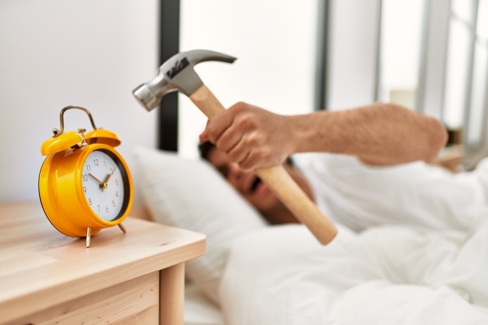 man turning off alarm clock using hammer lying on the bed at bedroom.