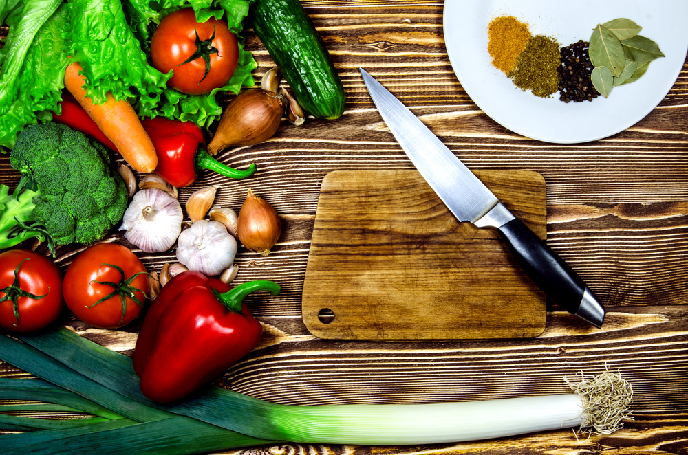 Red and green vegetables and greens lying on a wooden brown surface. Next to the vegetables is a small wooden plate and knife. On a white plate salad spices lie.