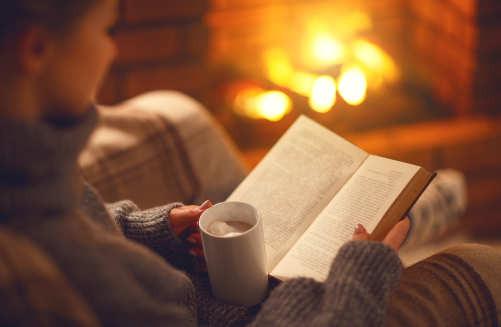 book and cup of coffee in hands of girl near fireplace