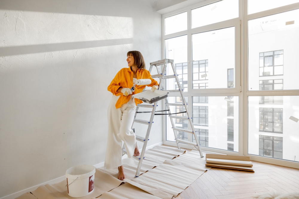 young woman making repairing in apartment, standing with paint roller near ladder in bright room of her new home