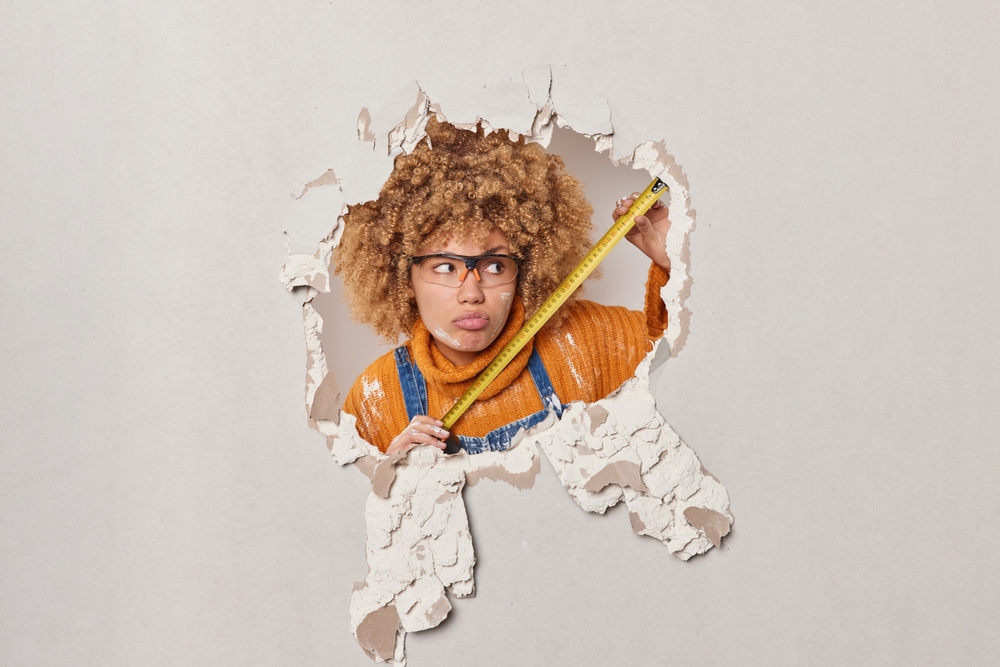 carpenter wears protective glasses and orange jumper holds measuring tape busy constructing focused aside looks through hole made in plaster wall.