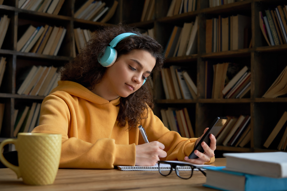 college student using smartphone watching video course, zoom calling making notes in workbook sit in library campus