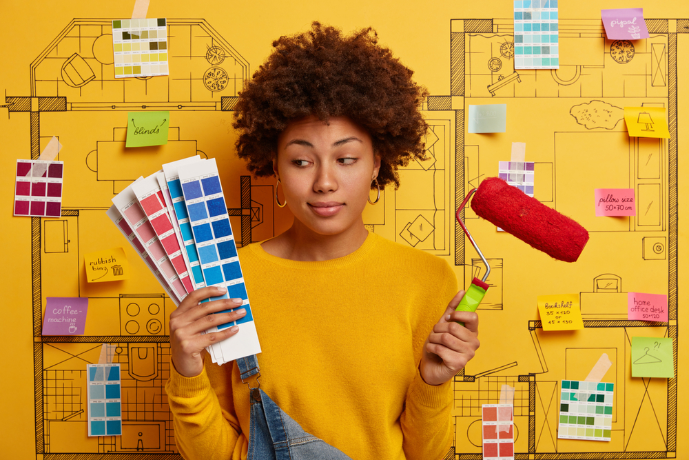 Thoughtful indecisive woman looks at color samples, holds paint roller, thinks about refurbishment of walls in new house, poses against sketch with sticky written notes.