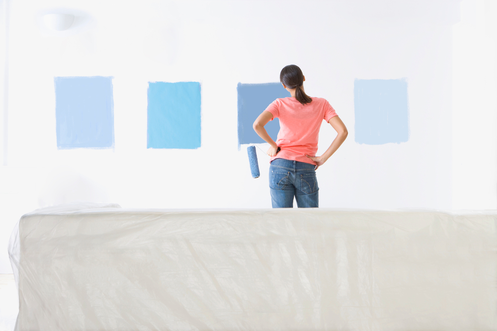 Woman holding a paint roller is looking at samples of blue paint on the living room wall to decide the right shade.
