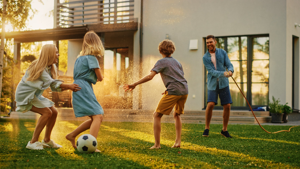 Happy Family of Four Playing with Garden Water Hose, Spraying Each Other. Mother, Father, Daughter and Son Have Fun Playing Games in the Backyard Lawn of Idyllic Suburban House on Sunny Summer Day