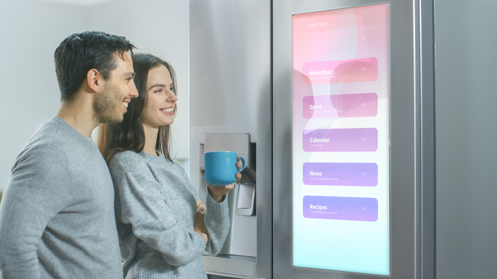 Young Couple Drink Morning Coffee in the Kitchen. They Check the Weather Forecast and a To Do List on a Smart Fridge at Home. Apartment is Bright and Cozy.