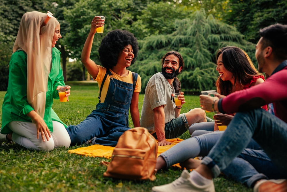 A group of diverse friends are sitting on the lawn of the public park in the city having drinks and a small party during the day.