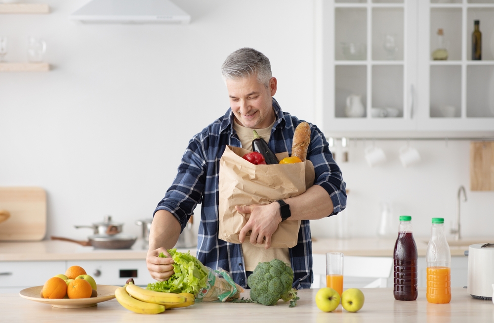 Mature man unpacking paper bag with fruits and vegetables ordered from internet shop, standing in kitchen interior, free space
