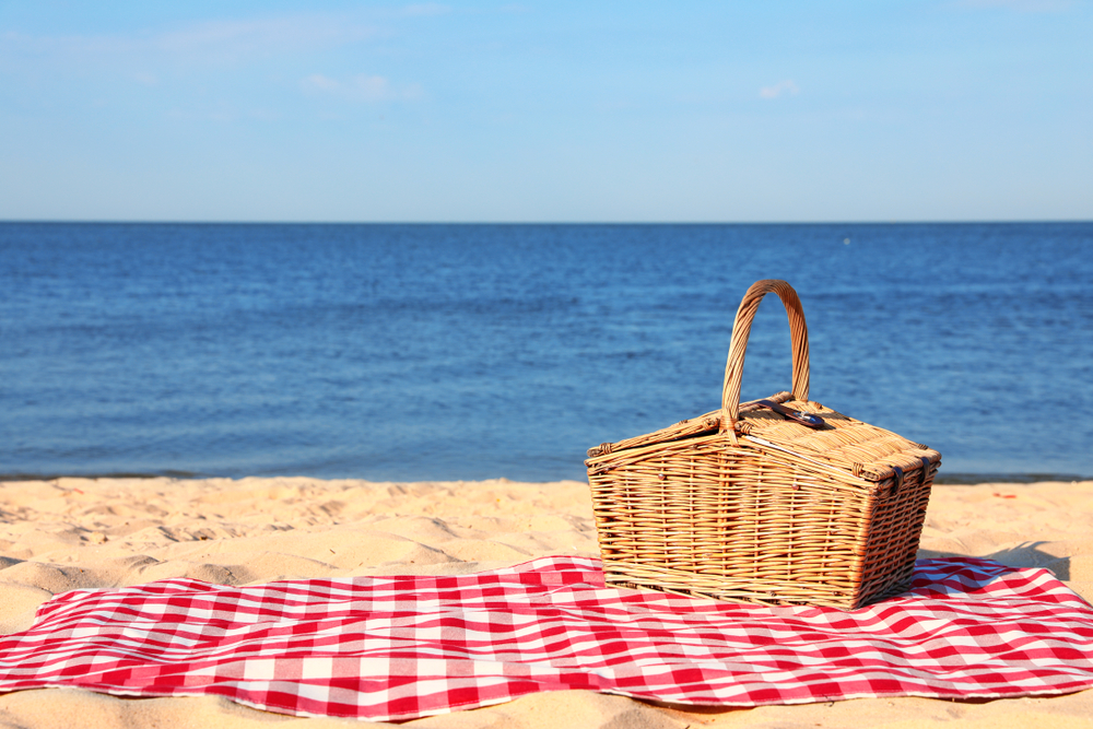 Checkered blanket with picnic basket on sunny beach. Space for text