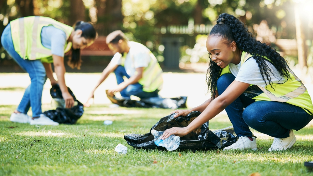 Volunteer people, cleaning park and community service for clean environment, recycling and pollution. Black woman happy to help group with plastic bottle and trash bag outdoor in green environment