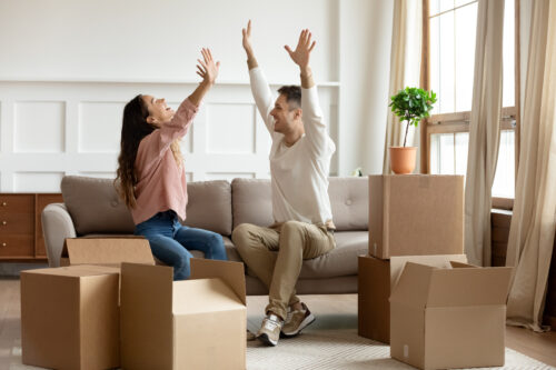 Happy young couple raising hands, sitting on couch in living room with cardboard boxes, celebrating moving day, family excited by relocation into first new house, buying real estate, mortgage